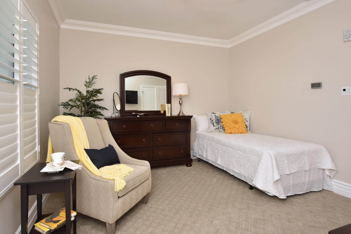 A cozy bedroom featuring a single bed, dresser with mirror, and armchair, complemented by a potted plant.