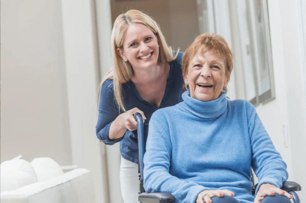 A caregiver happily assisting an elderly woman in a wheelchair indoors.