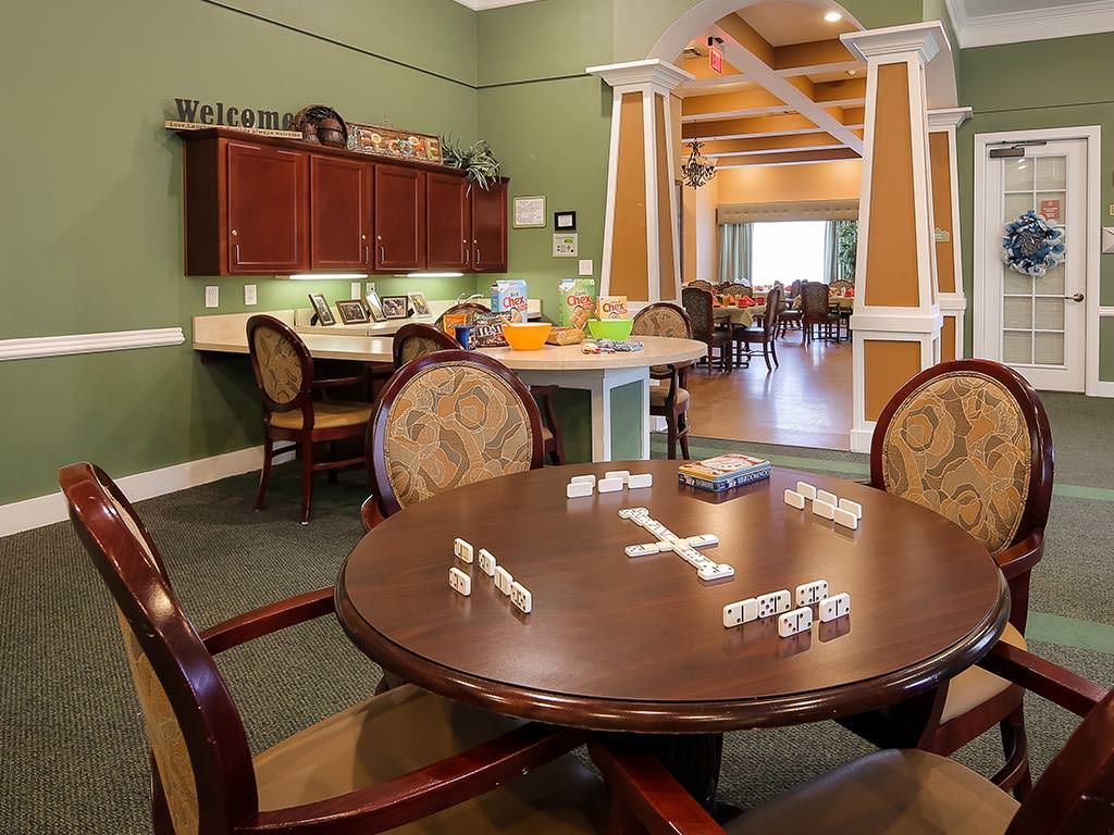 A cozy recreation room featuring a round table set up for a game of dominoes, with a kitchen area in the background.