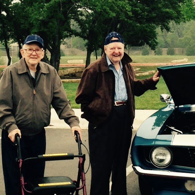 Two elderly men standing next to a vintage car in an outdoor setting.