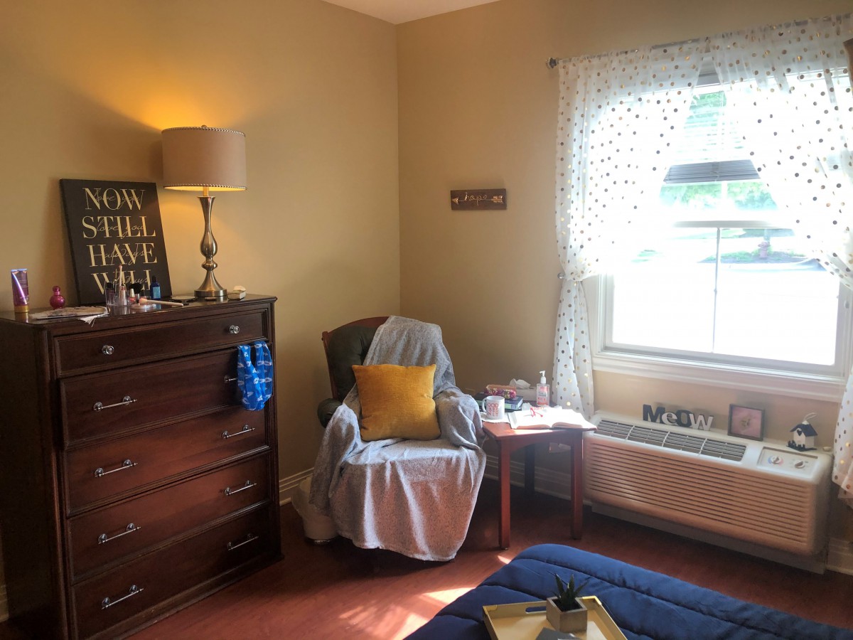 A cozy bedroom corner featuring an armchair with a yellow pillow, next to a window with sheer curtains and a wooden dresser.
