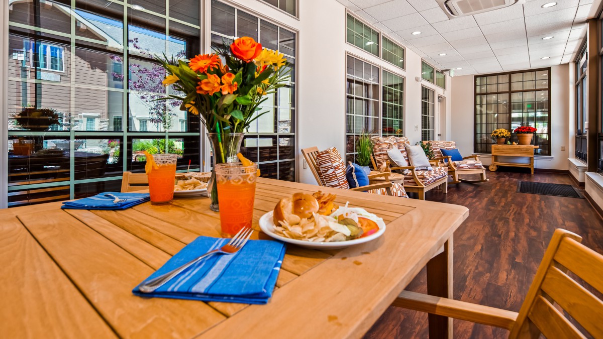 A cozy outdoor dining setup with a wooden table, vibrant flowers, and a plate of lunch accompanied by refreshing orange juice on a porch.