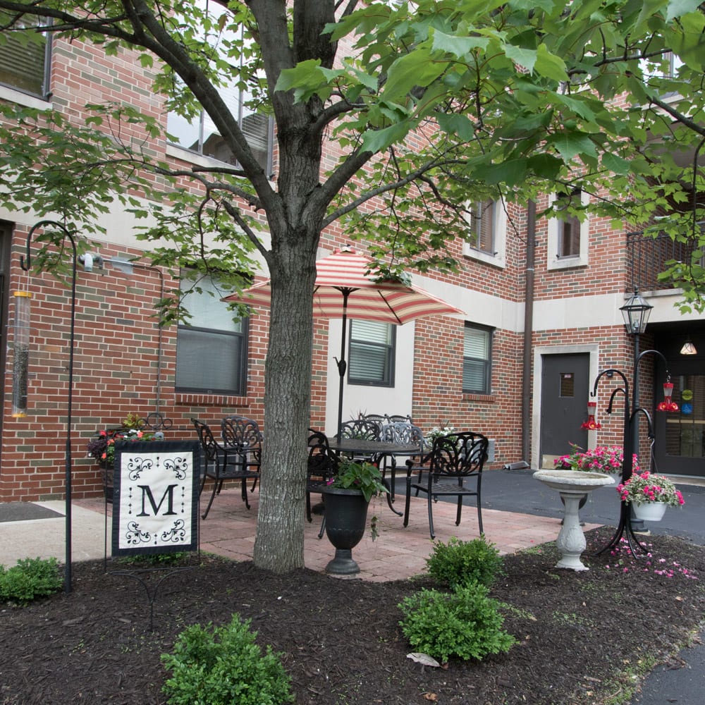 A cozy outdoor patio area with seating in front of a brick building, surrounded by greenery and featuring decorative signage.