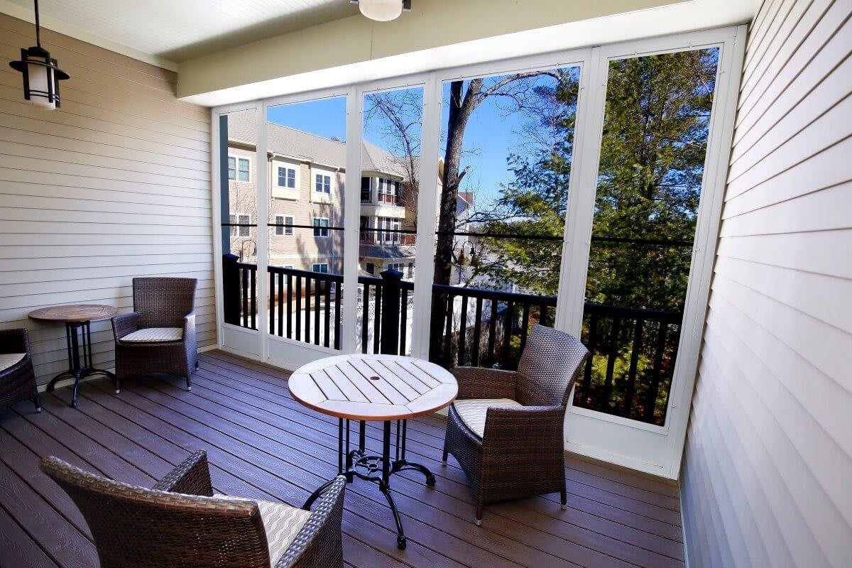 A cozy screened porch with outdoor furniture, featuring a round table and wicker chairs, overlooking trees and nearby buildings.