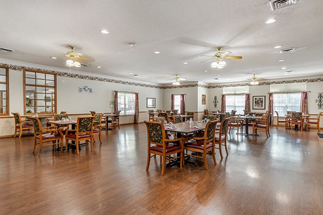 A spacious dining room with wooden floors and multiple tables and chairs, featuring ceiling fans for ventilation.