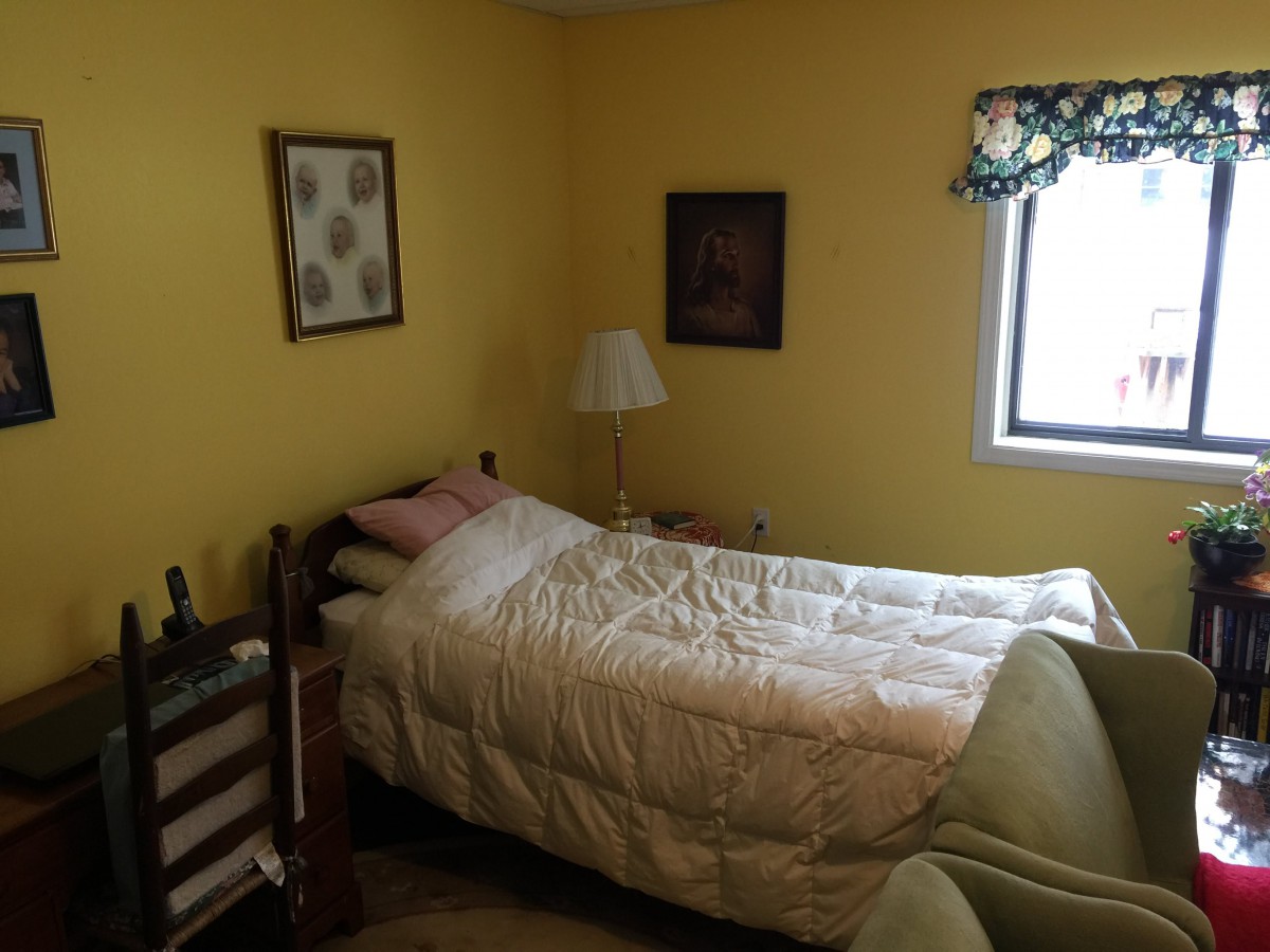 A cozy bedroom with yellow walls, featuring a single bed, artwork on the walls, a lamp, and a window with floral curtains.