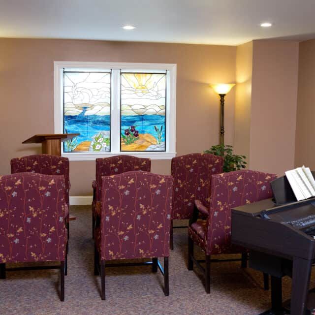 A small chapel interior with rows of chairs, a piano, lectern, and stained glass windows.