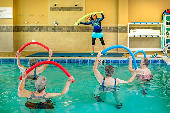 A group of seniors participating in a water aerobics class, using colorful noodles in an indoor swimming pool.