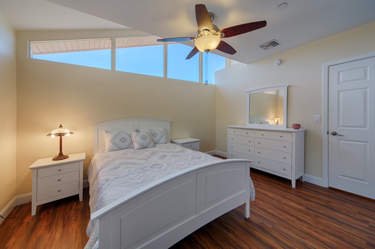 A cozy bedroom with a white bed, dresser, and wooden flooring, featuring a ceiling fan and ample natural light.