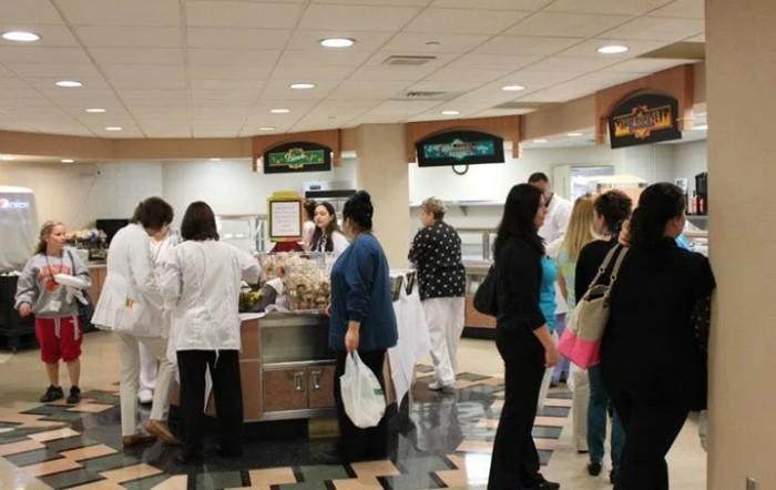 People in a cafeteria, including hospital staff and visitors, standing in line at the food service counter.