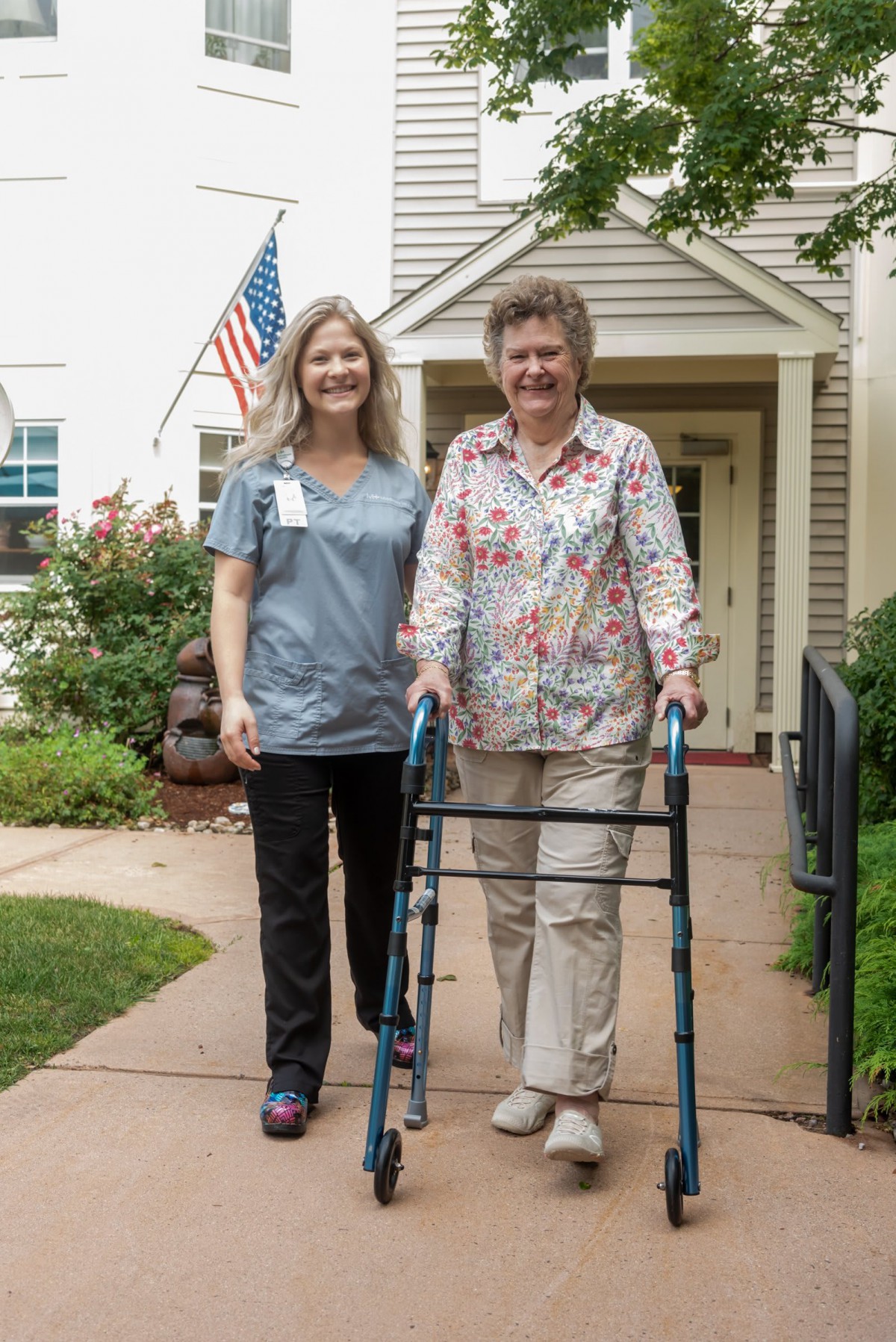 A nurse assists an elderly woman using a walker outside a residential home.
