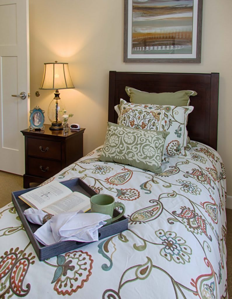 A cozy bedroom setting with a bed adorned with patterned pillows and a tray with a book and mug on top. A lamp on the nightstand adds warmth to the room.