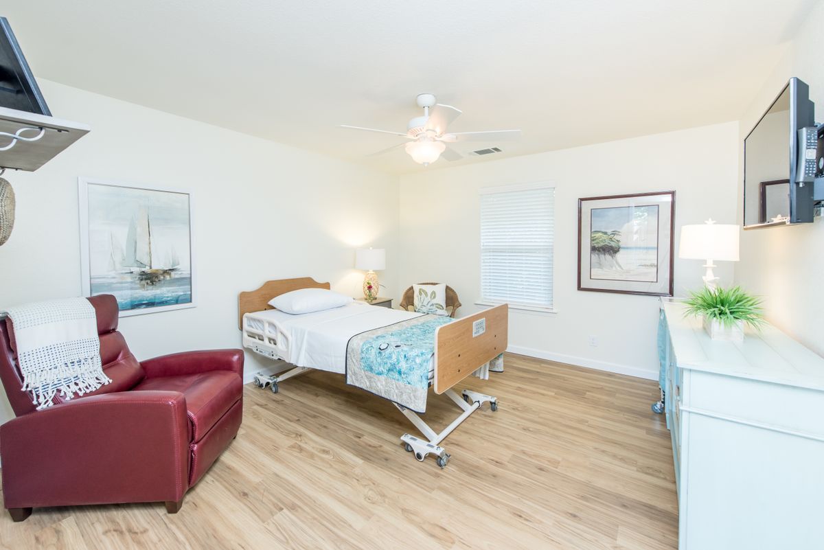 A clean and well-lit hospital room featuring a single adjustable bed, a red chair, framed artwork on the walls, and a wooden floor.