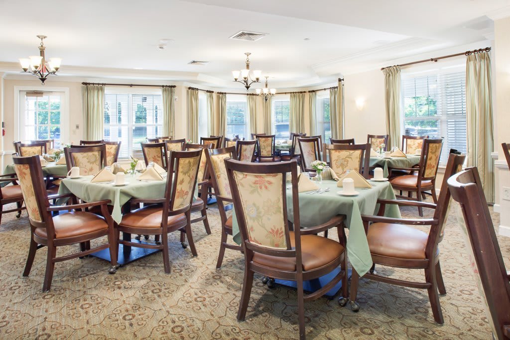 An elegant dining room with neatly arranged tables and chairs, featuring large windows and a central chandelier.