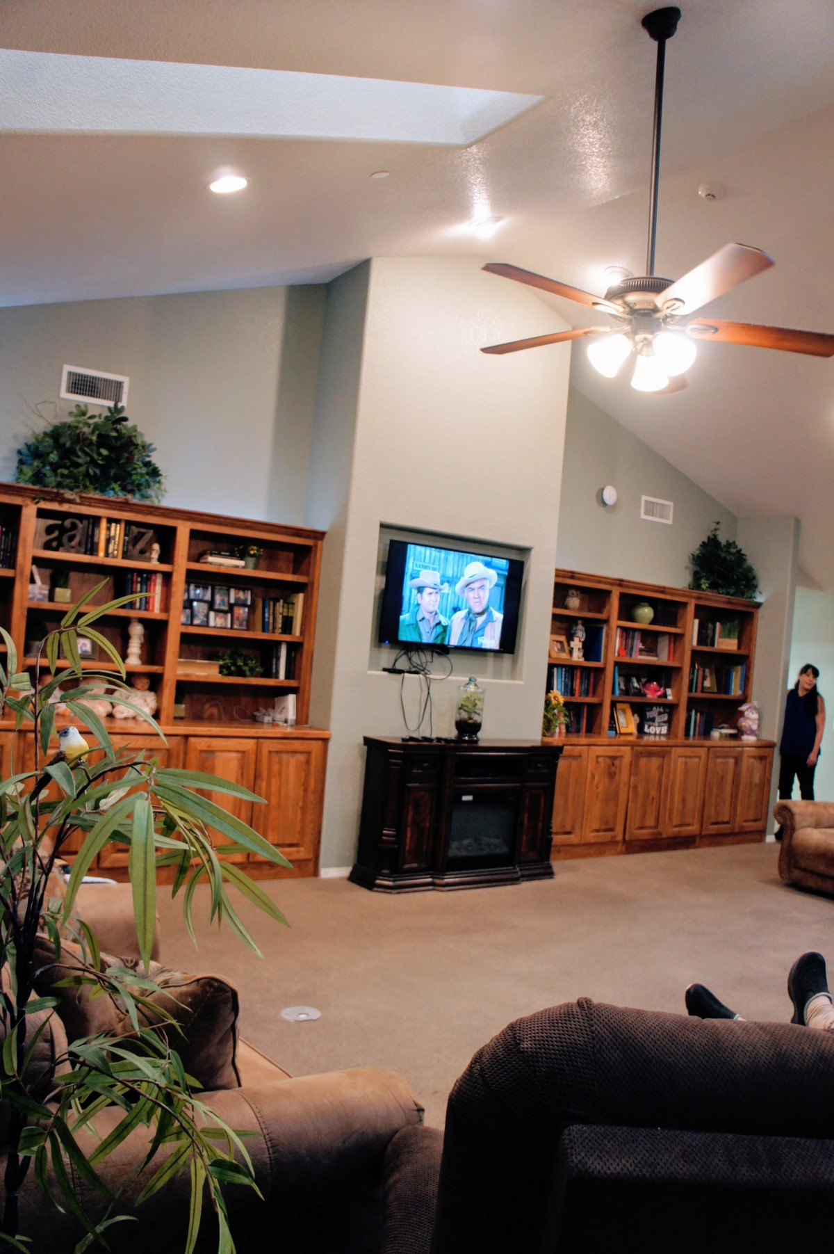 A cozy living room with wooden bookshelves, a mounted television, and a ceiling fan.