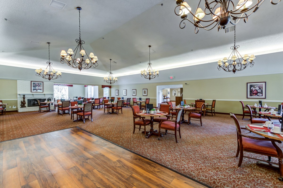 A spacious dining area with elegant chandeliers, tables, and chairs, featuring both carpeted and wood flooring.
