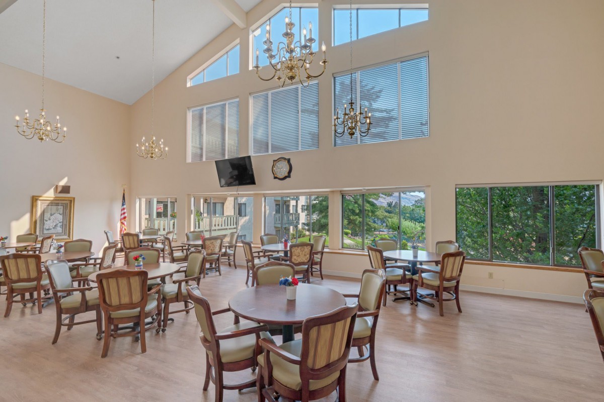 A spacious dining area with large windows, featuring multiple tables and chairs, chandeliers, and a wall-mounted television.