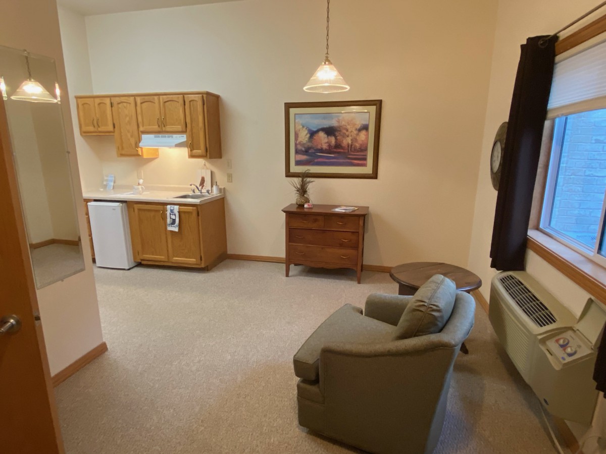 A cozy living space with a kitchenette, armchair, table, and wall painting.
