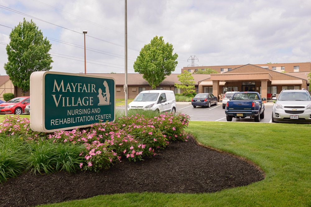 Entrance to Mayfair Village Nursing Home with visible sign, vehicles, and landscaped area.