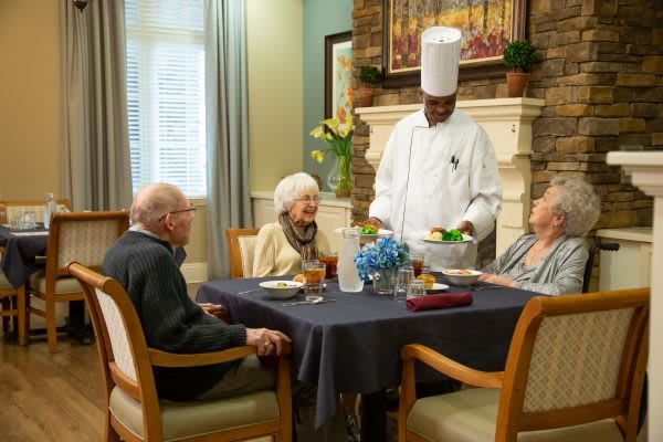 A chef serves a meal to elderly diners at a well-set table.