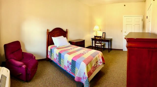 A cozy bedroom with a single bed adorned with a colorful quilt, alongside a small desk and lamp.