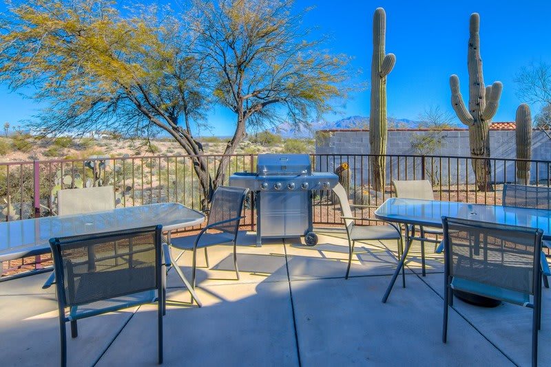 A sunny desert patio with a grill, tables, chairs, and cactus plants.