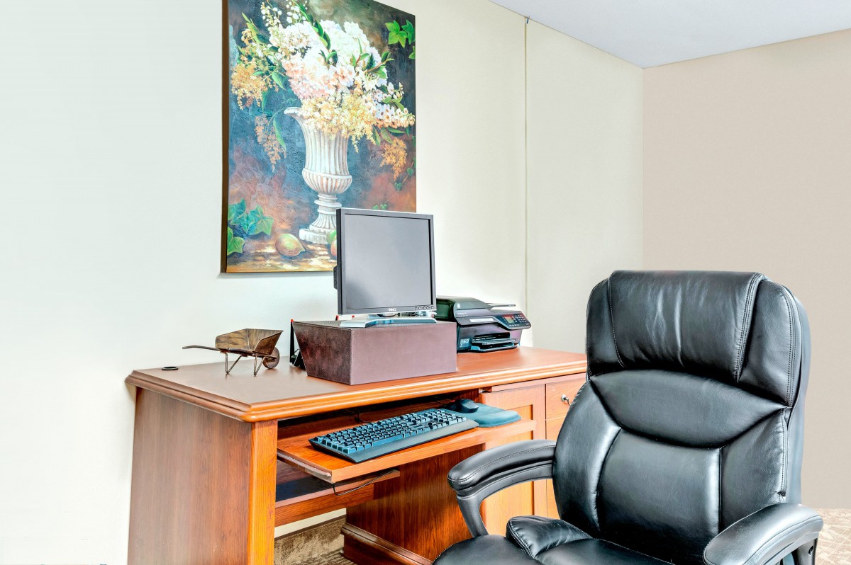 An office workspace featuring a wooden desk with a computer, keyboard, and office chair. A decorative painting hangs on the wall.