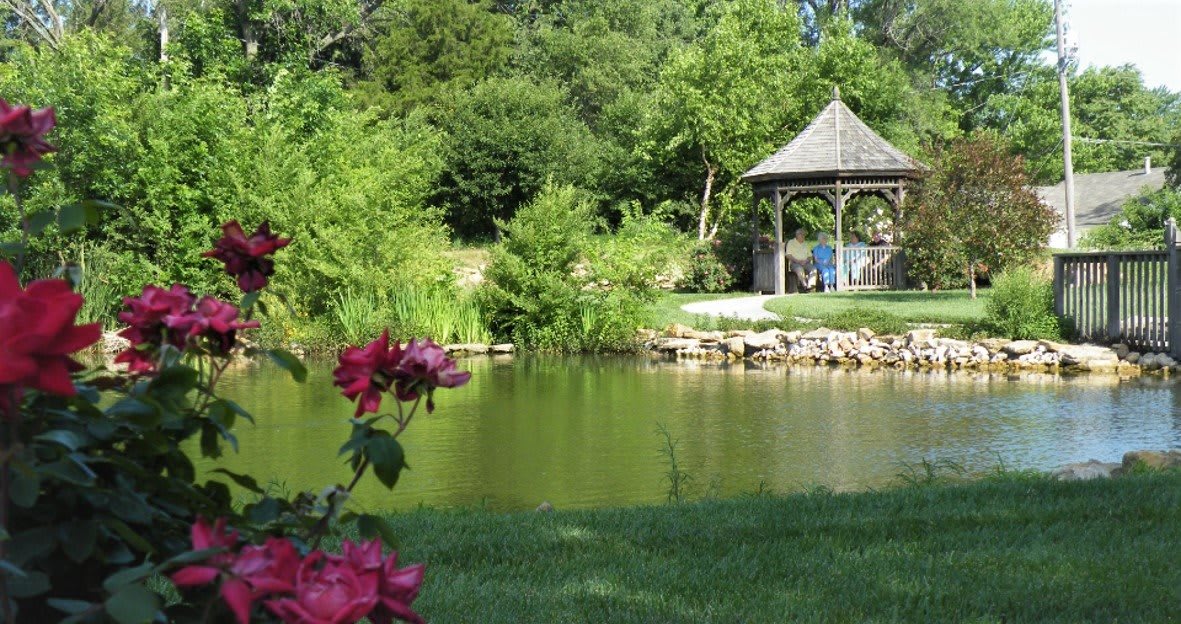 A peaceful garden scene with a pond and a gazebo surrounded by lush greenery and roses.