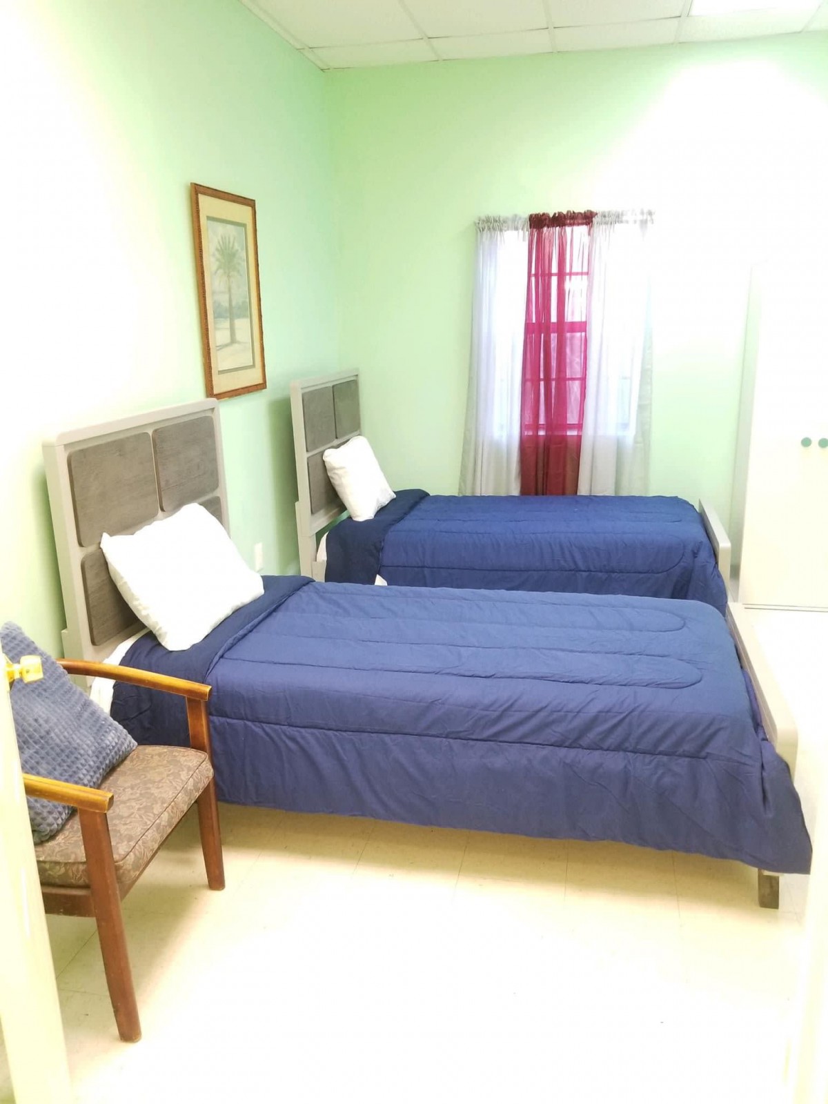 A tidy bedroom featuring two single beds with blue bedding, a window with curtains, a framed picture on the wall, and a chair by the bedside.