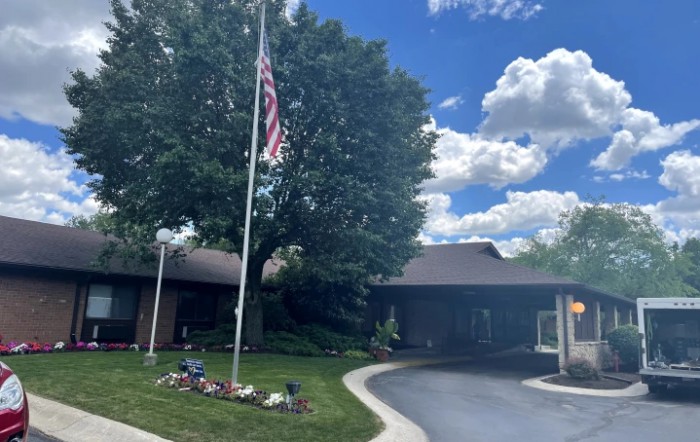 A residential house with a driveway and an American flag, surrounded by trees under a partly cloudy sky.
