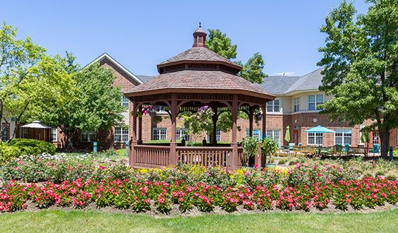 A wooden gazebo surrounded by vibrant flowers in a garden setting with buildings in the background.