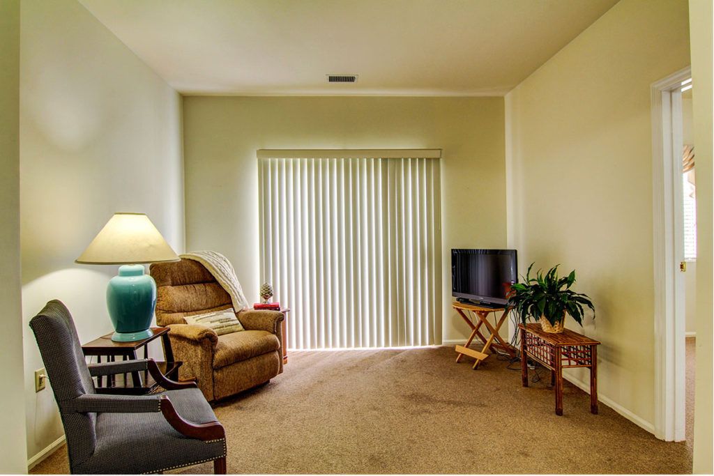 A cozy living room with a recliner chair, lamp, small television on a stand, and vertical blinds.