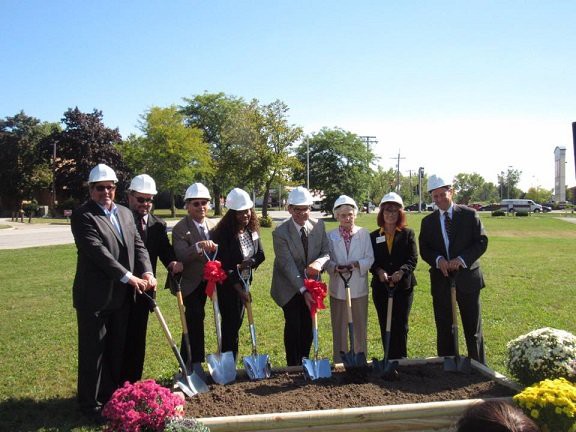 A group of people wearing hard hats participate in a groundbreaking ceremony outdoors, holding shovels and standing in front of a prepared plot of land.