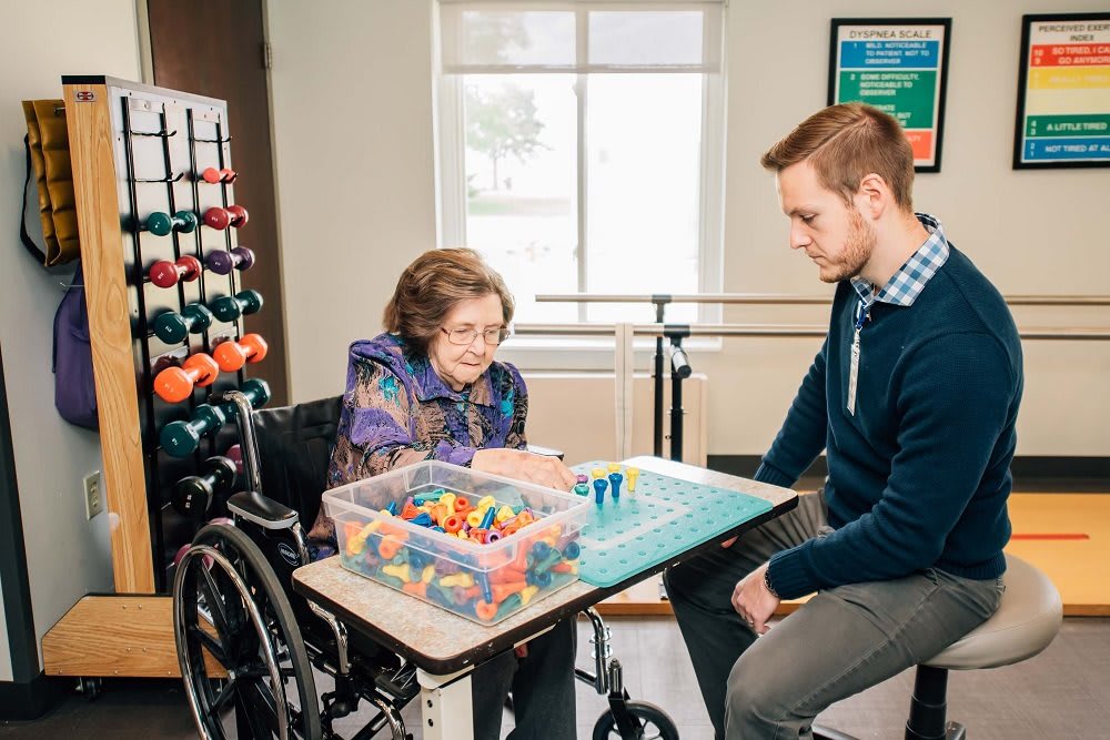 An elderly woman in a wheelchair is engaged in therapy activities with the assistance of a healthcare professional.