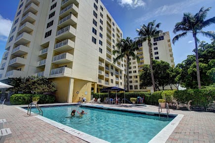 An outdoor swimming pool surrounded by apartment buildings and palm trees under a clear blue sky.