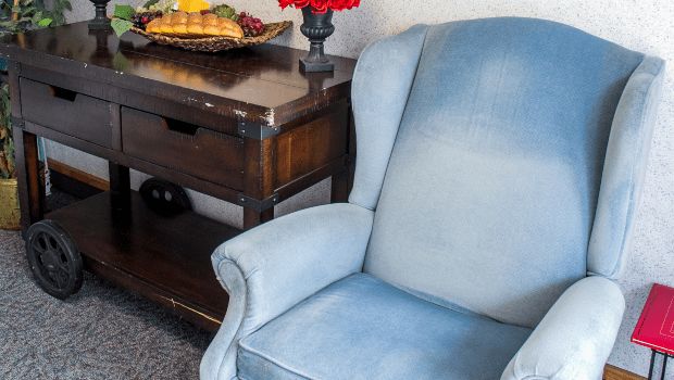 A cozy living room corner with a blue armchair beside a wooden table decorated with flowers and a fruit tray.