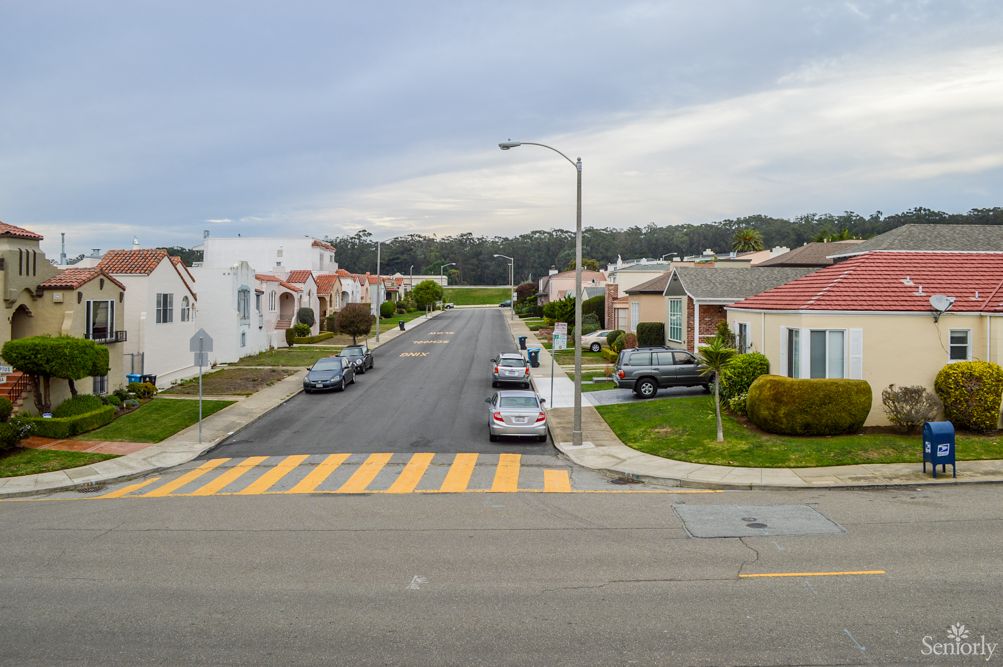 A quiet suburban street lined with houses and parked cars under an overcast sky.