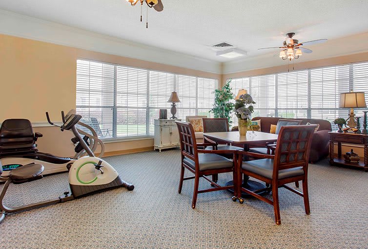 A well-lit room featuring an exercise bike, a table with chairs, and large windows offering ample natural light.
