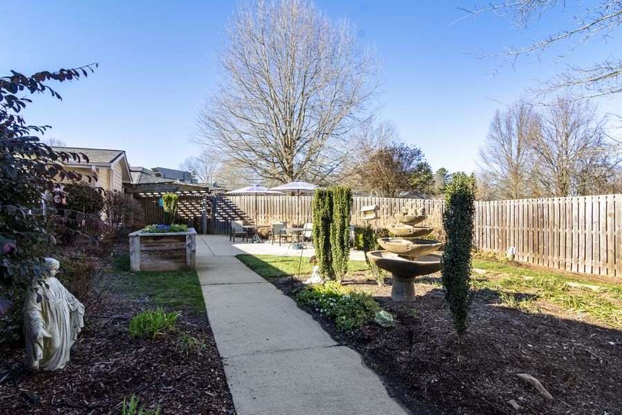 A serene backyard garden with a central concrete pathway surrounded by trees and plants under a clear blue sky.