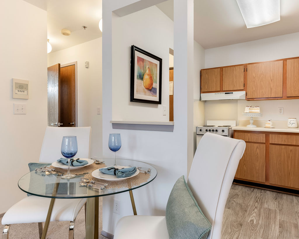 A modern apartment dining area with a glass table set for two, adjacent to a compact kitchen.