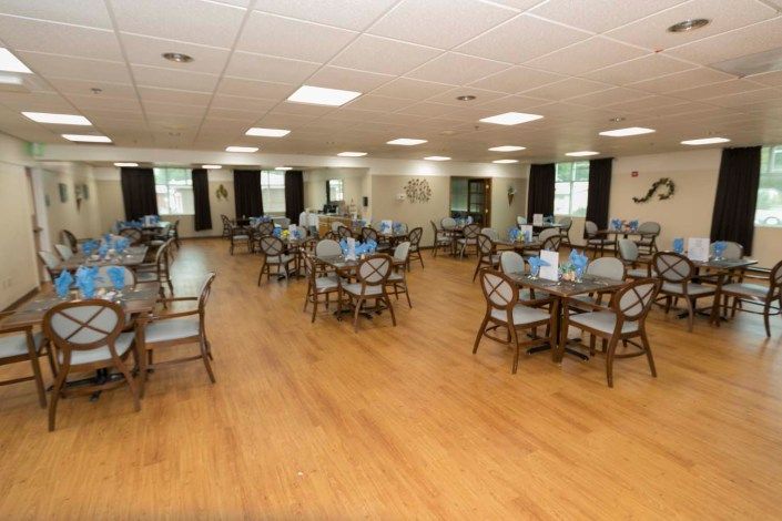 A spacious dining hall with wooden flooring, featuring multiple tables and chairs arranged neatly.
