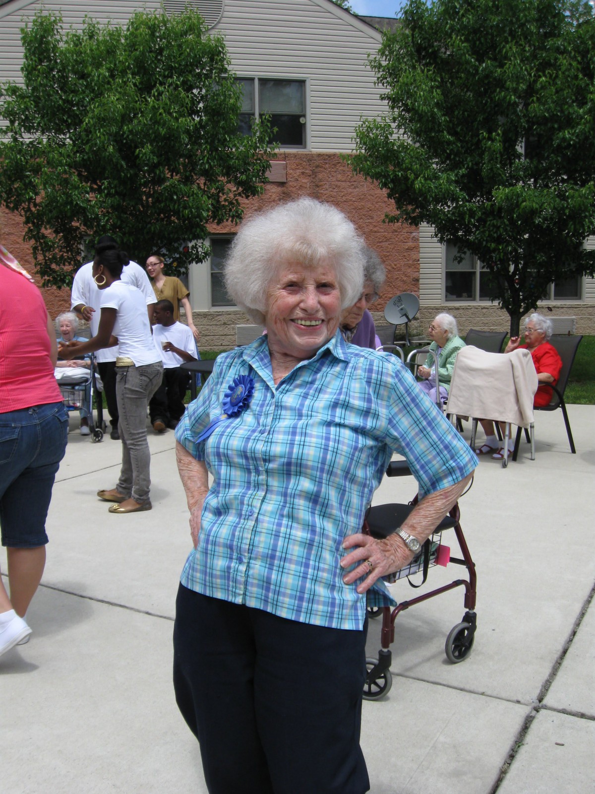 An elderly woman poses confidently at an outdoor gathering, with others socializing in the background.