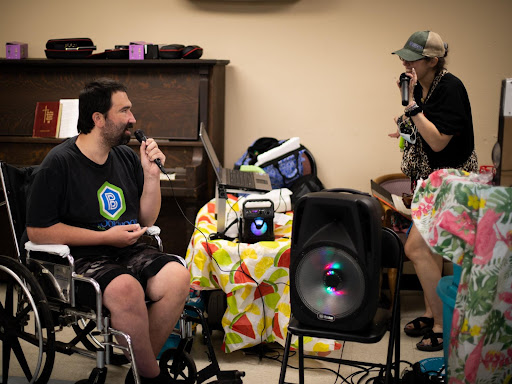 A lively indoor performance featuring a man in a wheelchair singing while another person looks on, surrounded by musical equipment.