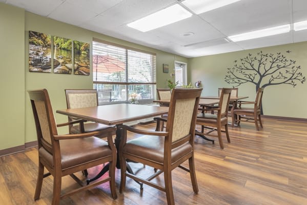 A bright dining room with wooden tables and chairs, featuring artwork on the walls and natural light from a large window.