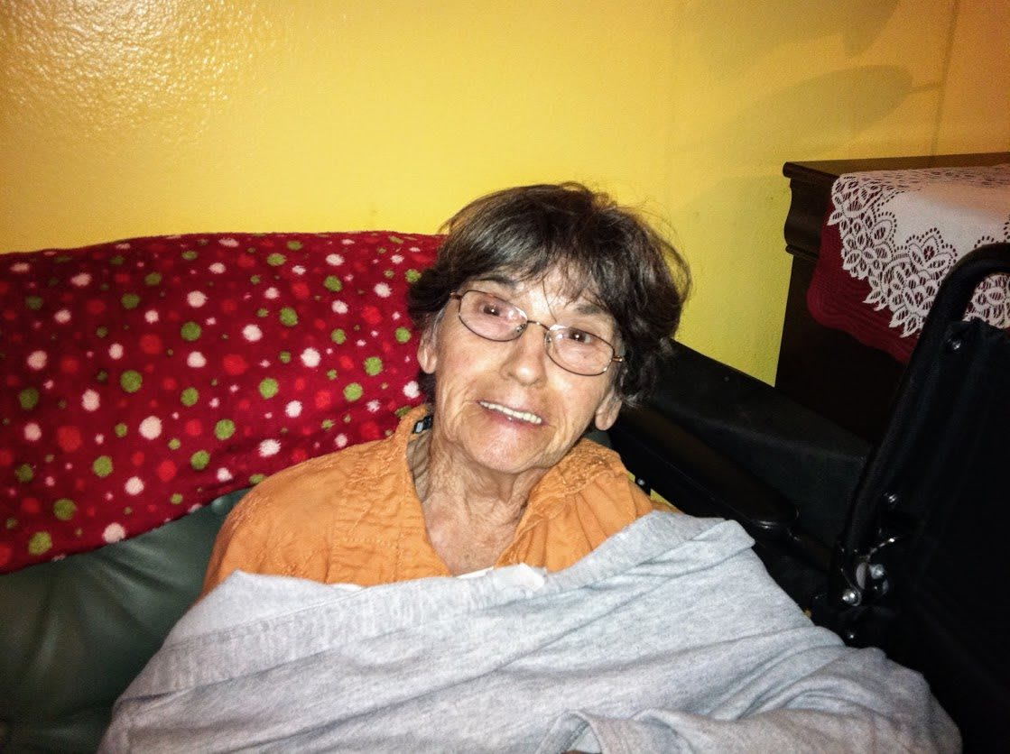 An elderly woman wearing glasses and smiling, wrapped in a blanket, seated indoors.