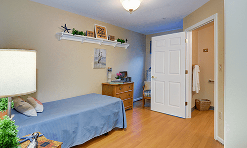 A cozy bedroom featuring a single bed with blue bedding, a wooden dresser, and a shelf above.