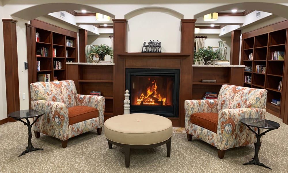 A cozy living room with a fireplace, two patterned chairs, and a round table, surrounded by bookshelves.