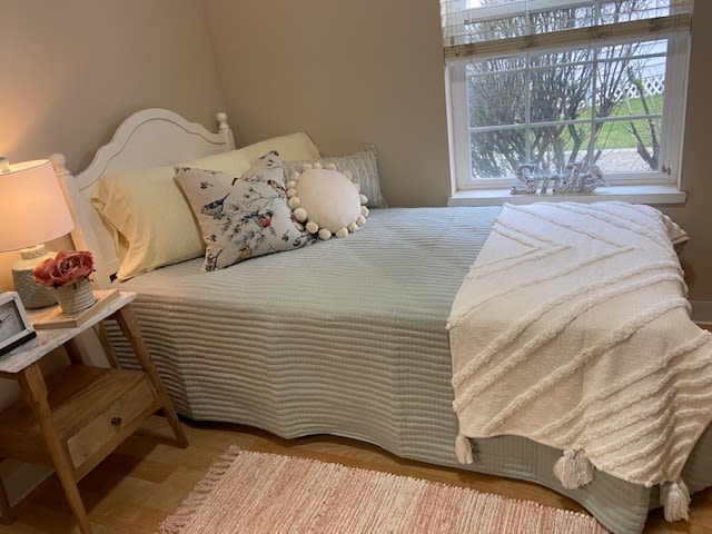 A cozy bedroom setup featuring a neatly made bed with decorative pillows and a blanket, situated by a window with soft natural light.