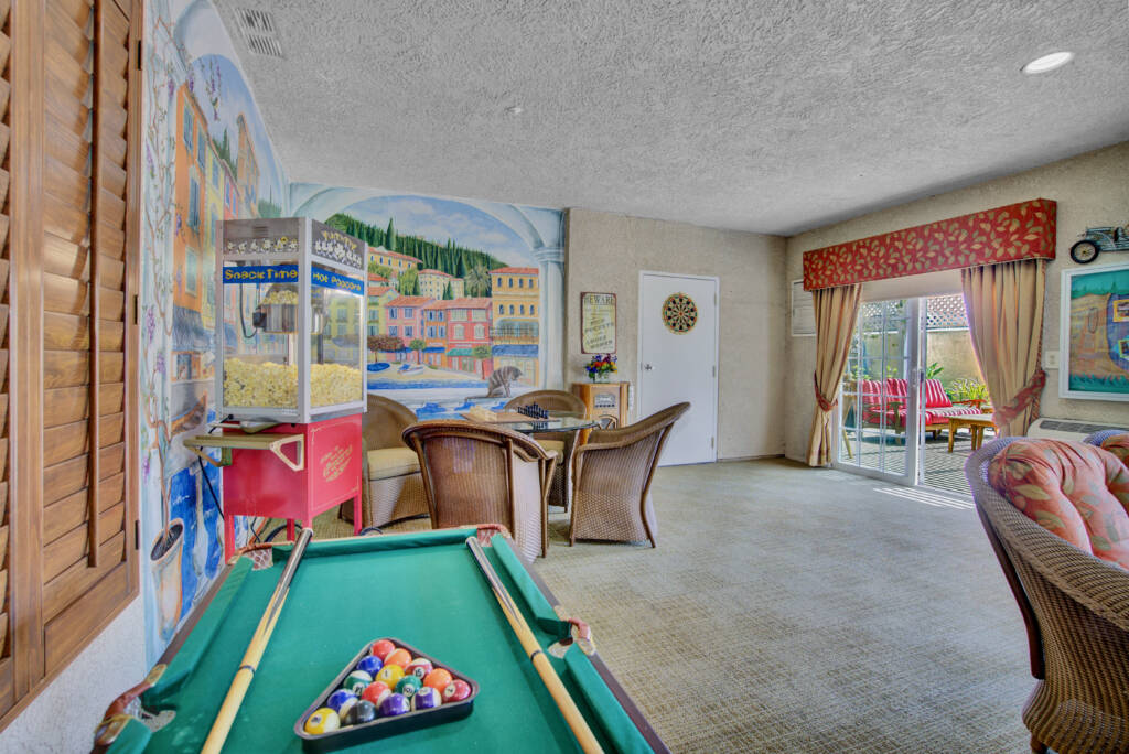 A colorful indoor game room featuring a pool table and a mural on the wall, with comfortable seating and large windows adorned with curtains.