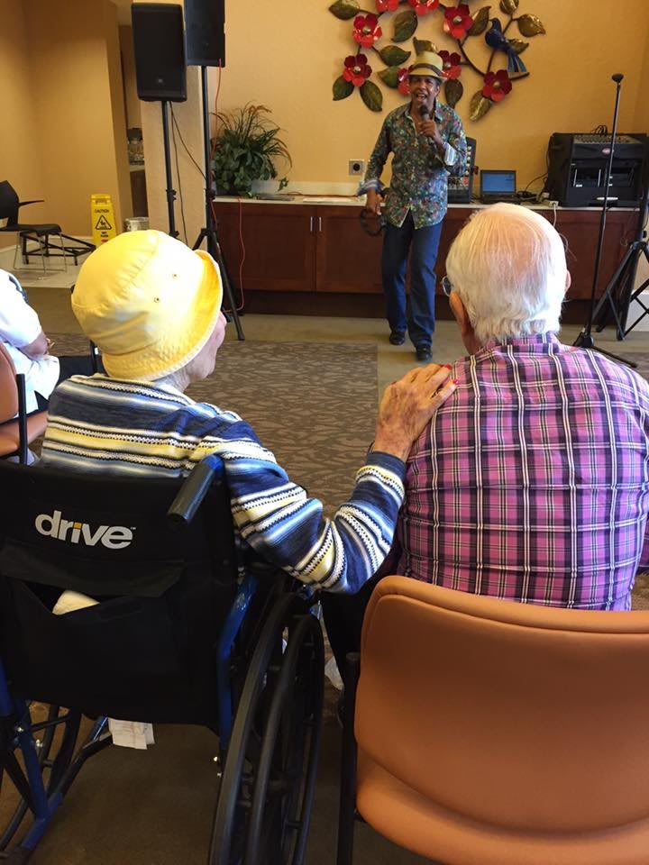 An elderly couple enjoying a moment together in a care facility, with one person seated in a wheelchair and gently resting their hand on the other's shoulder.